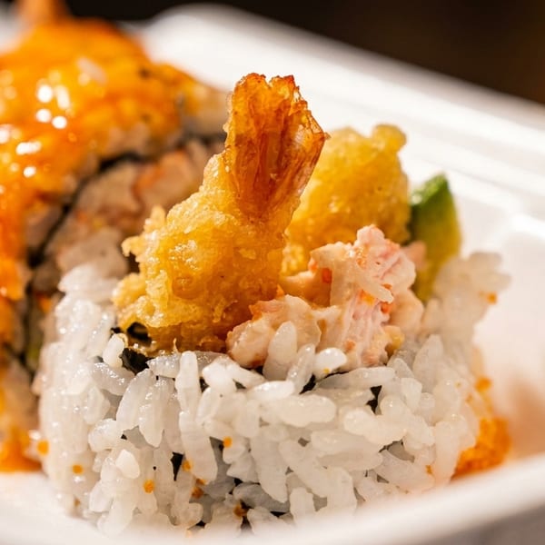 Close-up of sushi rolls with tempura shrimp, crab salad, and orange sauce in a white container.