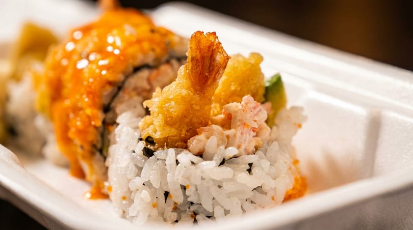 Close-up of sushi rolls with tempura shrimp, crab salad, and orange sauce in a white container.