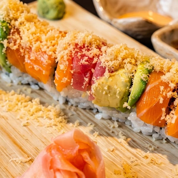 Colorful sushi roll with tuna, salmon, and avocado, topped with tempura flakes and served with ginger.