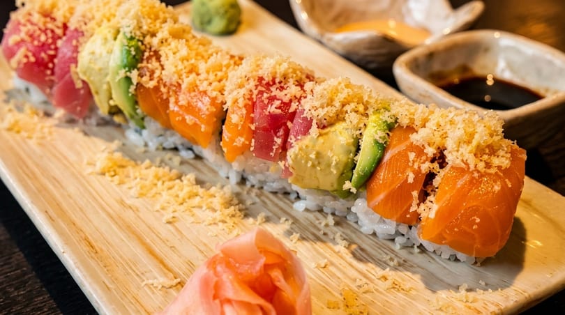 Colorful sushi roll with tuna, salmon, and avocado, topped with tempura flakes and served with ginger.