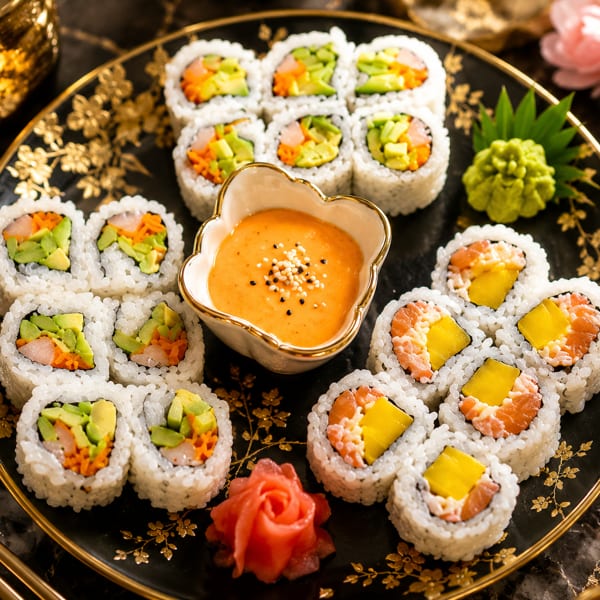 Assortment of sushi rolls with avocado, salmon, and mango, served with dipping sauce and ginger.
