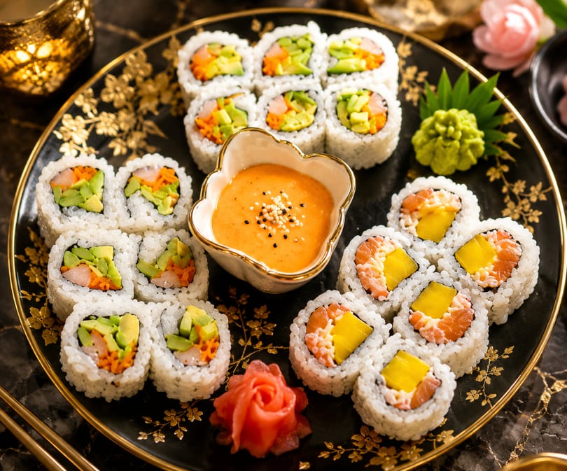 Assortment of sushi rolls with avocado, salmon, and mango, served with dipping sauce and ginger.