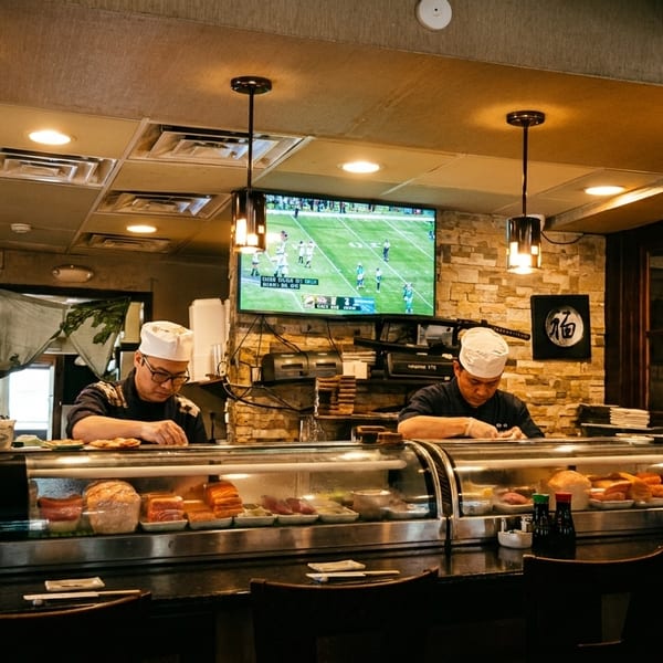 Two sushi chefs prepare food behind a counter with a football game playing on a TV in the background.