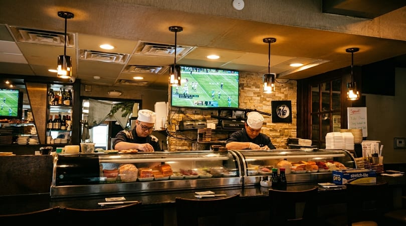 Two sushi chefs prepare food behind a counter with a football game playing on a TV in the background.