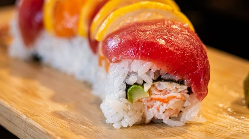 Close-up of a sushi roll with tuna, crab, avocado, and lemon slices on a wooden board.